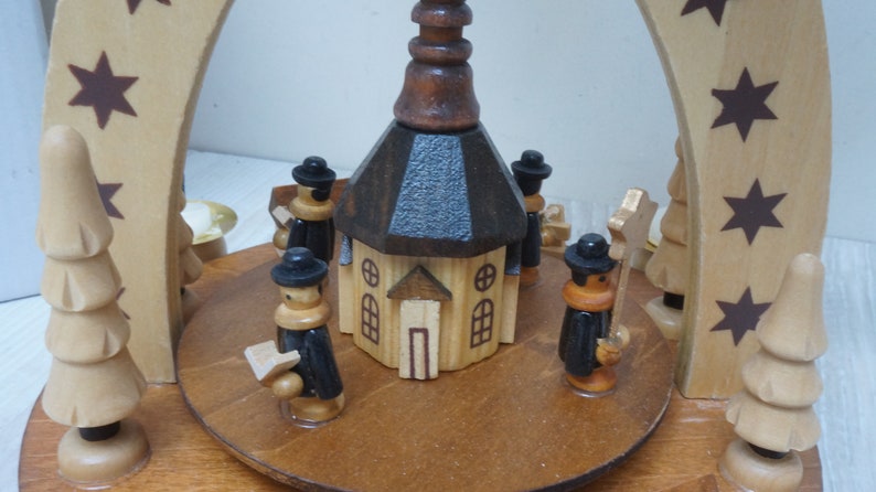 May include: A wooden Christmas pyramid with a church in the center and figures of people around it. The church has a brown roof and a blue roof. The figures are wearing black hats and coats. The pyramid is decorated with stars.