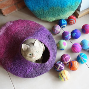 May include: A purple and pink felt cat bed with a white cat inside. The bed is surrounded by colorful felt balls.