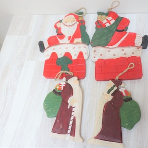 Op de afbeelding: Vier houten kerstversieringen met daarop Sinterklaas. Twee versieringen tonen Sinterklaas die een rode bakstenen schoorsteen afdaalt, terwijl de andere twee Sinterklaas tonen met een groene zak vol cadeaus. De versieringen zijn geschilderd in rood, wit, groen en bruin.