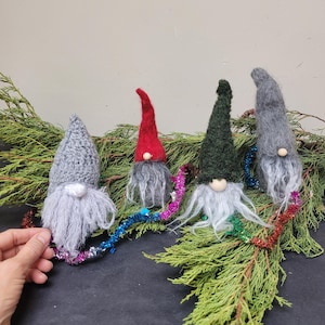 May include: Four handmade gnome ornaments with gray, red, and green pointed hats and long gray beards. Each gnome has a wooden nose and is decorated with colorful tinsel and placed on a bed of green foliage.