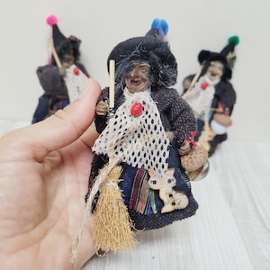 Puede incluir: Muñeca de bruja hecha a mano con sombrero negro, escoba y chal de encaje blanco. La muñeca tiene una cara detallada y lleva un abrigo oscuro. Un botón de madera con forma de gato está adherido al vestido de la muñeca.