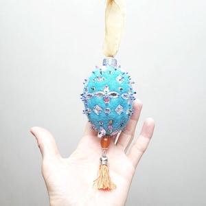 Könnte beinhalten: Ein türkisfarbener Weihnachtsbaumschmuck aus Filz mit einem Perlenmuster und einem Quasten. Der Schmuck ist mit Pailletten und Strasssteinen verziert.