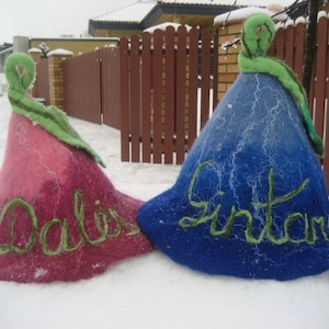 Puede incluir: Dos sombreros de fieltro con tallos verdes y nombres bordados. El sombrero rosa dice "Dalia" y el sombrero azul dice "Gintaras".