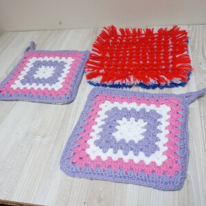 Könnte beinhalten: Drei gehäkelte Topflappen in verschiedenen Farben und Mustern. Einer ist rot mit weißen Fransen, einer ist lila, weiß und rosa, und der andere ist eine kleinere Version des lila, weißen und rosa Topflappens.