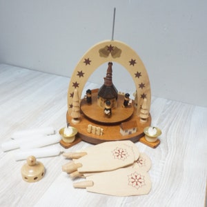 May include: A wooden Christmas pyramid with a brown church and four figures, surrounded by white candles, a base with a snowflake design, and a wooden star on top.
