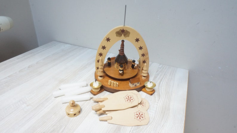 May include: A wooden Christmas pyramid with a brown church and four figures, surrounded by white candles, a base with a snowflake design, and a wooden star on top.