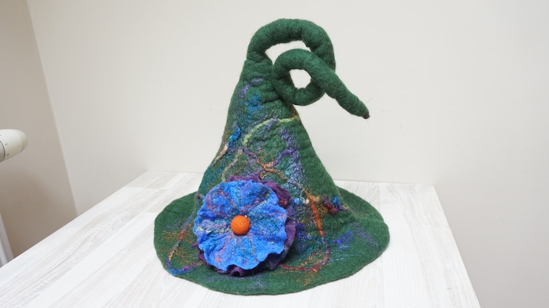 Op de afbeelding: Een groene vilten toverhoed met een gekrulde rand en een blauwe bloem met een oranje hart.