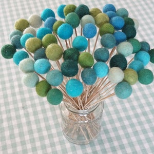 May include: A bouquet of felt balls in shades of blue and green, arranged in a clear glass vase. The felt balls are attached to wooden skewers.