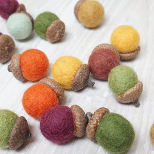 Op de afbeelding: Een verzameling van vilt eikel ornamenten in verschillende kleuren, waaronder rood, oranje, geel, groen, bruin en paars. De eikels zijn allemaal handgemaakt en hebben een rustieke, natuurlijke uitstraling.