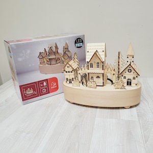Könnte beinhalten: Eine Weihnachtsdorf-Szene aus Holz mit drehbarer Basis, bestehend aus einer Kirche, Häusern und Bäumen. Das Dorf wird von 9 LED-Lichtern beleuchtet und trägt auf der Verpackung den Schriftzug "LED 9x". Die Verpackung trägt außerdem den Hinweis "Indoor".
