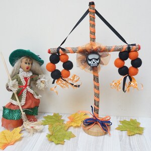 Może przedstawiać: Dekoracja halloweenowa z drewnianym krzyżem ozdobionym pomarańczową i czarną wstążką, ozdobami z filcowych kulek i zawieszką w kształcie czaszki. W pobliżu stoi lalka czarownicy w zielonym kapeluszu i z miotłą.
