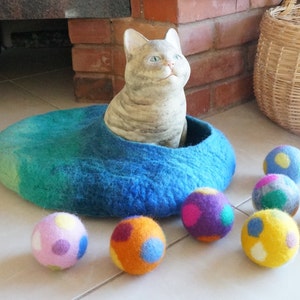 May include: A blue felt cat bed with a white cat figurine inside. The bed is surrounded by colorful felt balls with polka dots.