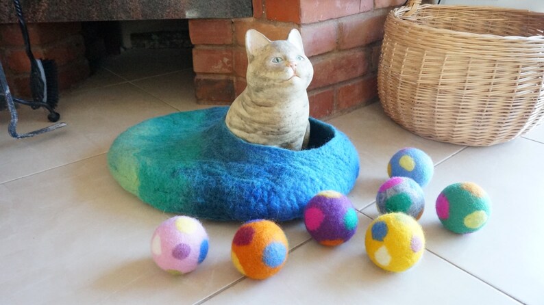 May include: A blue felt cat bed with a white cat figurine inside. The bed is surrounded by colorful felt balls with polka dots.