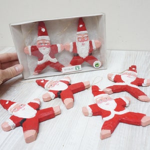 Op de afbeelding: Vijf houten kerstversieringen in de vorm van kerstmannen, geschilderd in rood en wit. De versieringen zijn stervormig en hebben een touwtje om ze op te hangen. De versieringen zitten in een doos met de tekst "81108.47" erop.