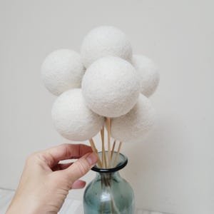 Könnte beinhalten: Ein Strauß weißer Wollkugeln auf Holzstäbchen in einer blauen Glasvase.