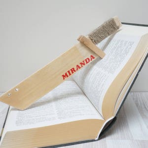 Op de afbeelding: Een houten bladwijzer in de vorm van een zwaard met het woord "MIRANDA" erop gedrukt in rode letters. De bladwijzer is geplaatst in een open boek.