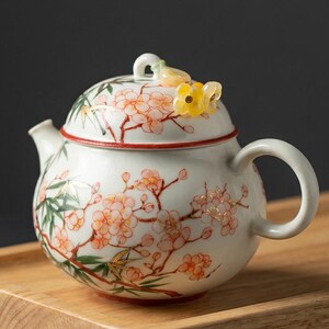 Pode incluir: Um bule de cerâmica branca com um design pintado à mão de flores de cerejeira cor-de-rosa e caules de bambu verdes. A tampa é rematada com um pequeno ornamento floral amarelo translúcido. O bule tem uma pega curva e um bico curto e cónico.