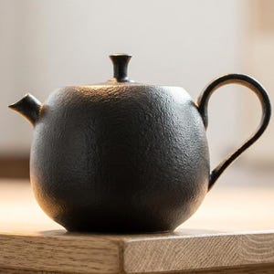 Puede incluir: Una tetera negra mate con cuerpo redondeado, pico corto, asa curva y una tapa pequeña con pomo. La superficie de la tetera tiene un acabado texturizado y granulado. La tetera se muestra de perfil, dejando ver su forma circular característica.