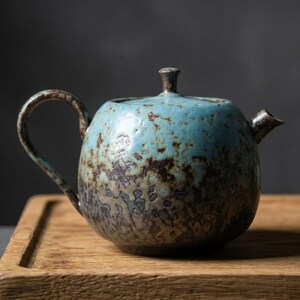 Op de afbeelding: Een ronde keramische theepot met een getextureerd oppervlak met een verloop van helder turkooisblauw aan de bovenkant dat overgaat in een donker, gevlekt bruin aan de basis, compleet met bijpassend deksel, gebogen handvat en kleine tuit.