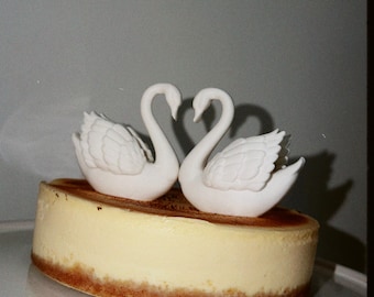 May include: A round cheesecake with a golden-brown crust and a smooth, pale yellow top, topped with two white swan figurines facing each other. The swans have detailed, textured wings and long, curved necks that form a heart shape between them.