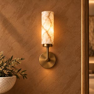 May include: A wall-mounted light fixture featuring a cylindrical shade with a white and brown marble pattern. The lamp is supported by a circular brass-toned metal base and a vertical arm, emitting a warm glow from within the shade.