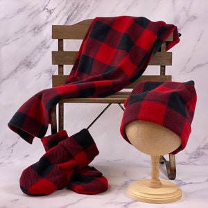 May include: Red and black buffalo check fleece blanket, hat, and booties. The blanket is draped over a wooden bench. The hat is on a wooden stand. The booties are on the floor.