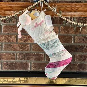 May include: A quilted white Christmas stocking featuring the name Morgan embroidered in pink script. The stocking is decorated with horizontal fabric strips in floral and lace patterns, and includes a small plush lamb and yellow toy inside.