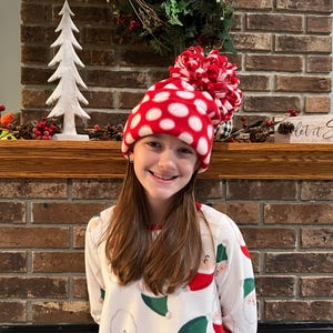 May include: A young person wearing a red and white polka dot fleece hat with a white pom pom on top. The hat is sitting on their head and they are smiling. The person is wearing a white shirt with a Christmas pattern. The background is a brick fireplace with a wooden mantel. There is a small white Christmas tree on the mantel and a sign that says "Let it Snow".