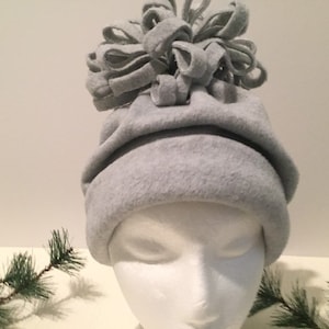 May include: A light gray fleece beanie hat featuring a wide folded brim and a large, decorative pom-pom on top constructed from multiple loops of the same gray fleece fabric. The hat is displayed on a plain white mannequin head form.