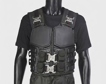 May include: A black tactical vest featuring padded chest panels, multiple integrated storage pouches, and silver-toned metal quick-release buckles on the shoulders and front closure. The vest is worn over a plain black short-sleeved t-shirt.