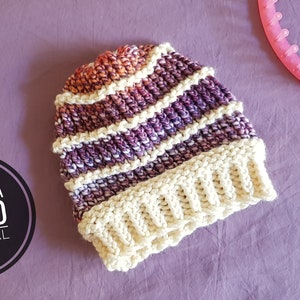 May include: A striped crochet hat with a cream colored brim. The hat is made with a gradient yarn that transitions from orange to purple. The hat is lying on a purple surface. Includes a video tutorial.