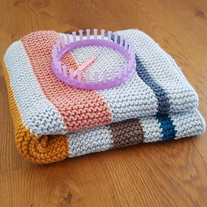 May include: A folded, striped knitted blanket in shades of gray, peach, yellow, and brown, with a purple knitting loom and hook on top. The blanket is made of thick yarn and has a textured appearance. The loom is circular.