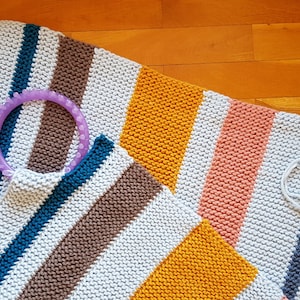 May include: Hand-knitted rectangular and square blankets with striped patterns in gray, brown, teal, orange, and pink. A purple knitting loom is placed on top of the blankets. The blankets are on a wooden surface.