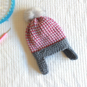 May include: A handmade winter hat with a gray brim and ear flaps, transitioning to a red, white, and gray patterned crown, topped with a fluffy gray pom-pom. A pink crochet hook, a blue loom, and a measuring tape are also visible.