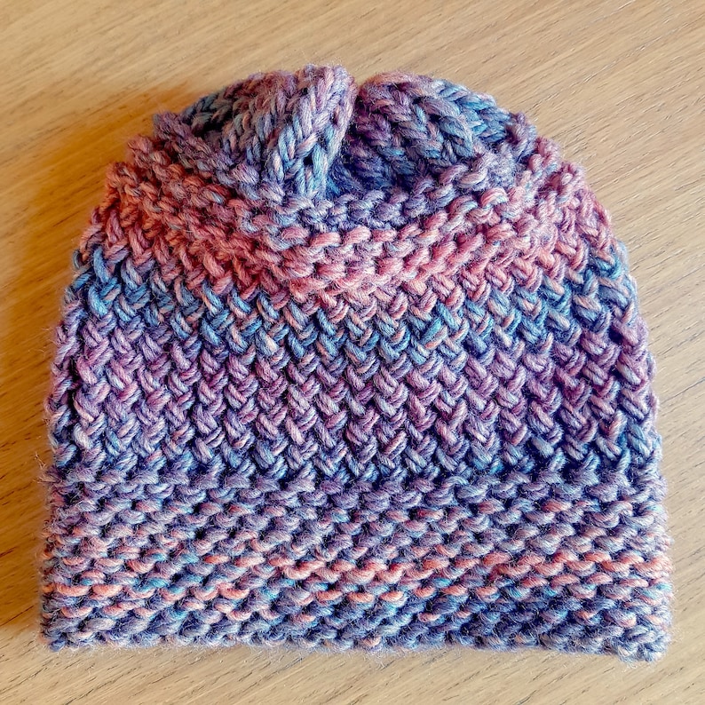 May include: A knitted hat in shades of pink, blue, and purple. The hat has a textured stitch pattern and a slouchy fit.
