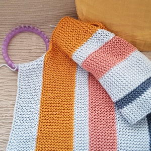 May include: A handmade knitted blanket with orange, pink, and gray stripes. A ball of light gray yarn and a purple knitting loom are also visible. The blanket is folded and ready to use.