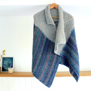 Puede incluir: Un chal de punto gris y azul con un patrón diagonal. El chal está colgado de una percha de madera contra una pared blanca.
