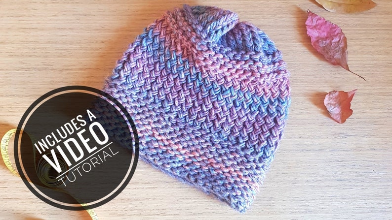 May include: A purple, pink, and blue knitted hat with a textured stitch pattern. The hat is on a wooden surface with a yellow measuring tape and fall leaves. The image includes the text "Includes a Video Tutorial".