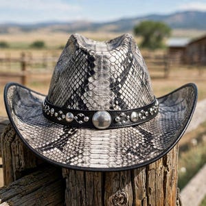 Op de afbeelding: Een cowboyhoed met een grijs en zwart slangenleerpatroon en een zwarte leren band. De band is versierd met zilverkleurige metalen studs en een centrale ronde concho. De rand heeft een zwarte afwerking en een gebogen, gestructureerde vorm.