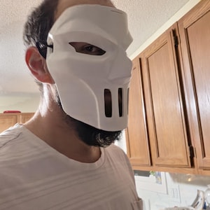 May include: A white, rigid, full-face mask featuring angular contours, almond-shaped eye openings, and a prominent nose bridge. The lower section includes vertical ventilation slits over the mouth area, secured by a black elastic strap.