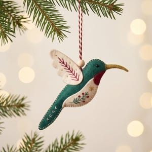 May include: A felt hummingbird ornament featuring a deep green body, cream-colored wings with red stitching, and a red throat patch. The bird has a long, pointed beak and is decorated with embroidered floral motifs. It hangs from a red and white string.