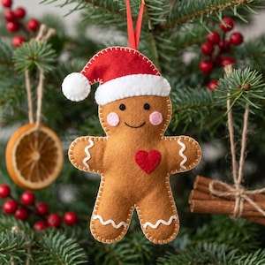 May include: A felt gingerbread person ornament featuring a red Santa hat with a white pom-pom, pink cheeks, a stitched smile, and a red heart on the chest. The figure has white stitched icing details on the arms and legs and hangs from a red ribbon.