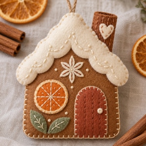 Peut inclure: Une décoration en feutrine en forme de maison en pain d'épices avec une base marron, un toit festonné crème brodé, une étoile centrale, une applique en forme de tranche d'orange, une porte cintrée rouge foncé et une cheminée avec un cœur.