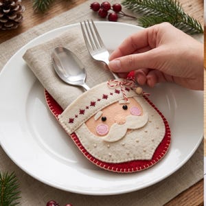 May include: A felt Santa Claus cutlery holder in red and cream, featuring a stitched face with pink cheeks and a white beard. The pouch contains a beige linen napkin, a silver spoon, and a silver fork, tied with a red and white string.