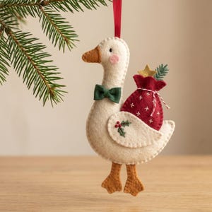 May include: A felt goose ornament in cream with a brown beak and feet, wearing a dark green bow tie. The goose carries a red sack with white cross-stitch details, a gold star, and a green sprig on its back. A small holly leaf is stitched on the wing.