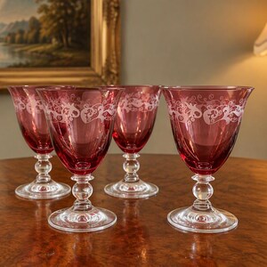 May include: A set of four cranberry-coloured glass goblets with clear stems and bases. Each goblet features a wide, rounded bowl with a delicate, etched white floral scroll pattern encircling the upper rim of the deep red glass.