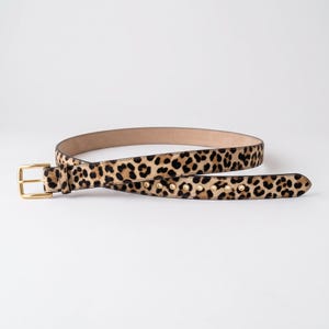 Peut inclure: Une ceinture avec un motif imprimé léopard beige et noir et une surface texturée. La ceinture comprend une boucle en métal doré poli et une série d'œillets en métal doré le long de la sangle, avec une doublure intérieure beige clair.