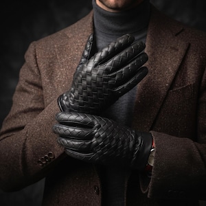 Könnte beinhalten: Ein Paar schwarze Lederhandschuhe mit einem komplexen Flechtmuster auf dem Handrücken und den Fingern. Die Handschuhe verfügen über ein glattes, einfarbiges Lederbündchen am Handgelenk und ein strukturiertes Design, das die gesamte Hand bedeckt.