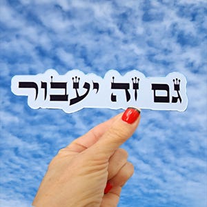 May include: A white die-cut sticker featuring black Hebrew text that reads "גם זה יעבור" with small crown icons integrated into the lettering. The sticker has a smooth, contoured edge and is presented against a bright blue sky with white clouds.