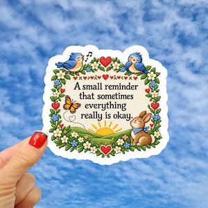May include: A die-cut sticker featuring a cross-stitch style design with a floral border, two blue birds, a butterfly, and a brown rabbit. The text reads, A small reminder that sometimes everything really is okay, centered above a rising sun.